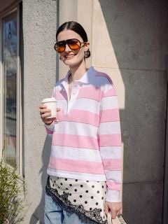 Eine lächelnde Frau mit Sonnenbrille hält eine Kaffeetasse, trägt ein rosa-weiß gestreiftes Hemd und einen gepunkteten Schal und steht draußen an einem Gebäude.