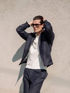 Ein Mann, der eine Sonnenbrille, eine marineblaue Jacke und eine dazu passende Hose trägt, steht mit den Händen auf dem Kopf vor einer hellen, strukturierten Wand.