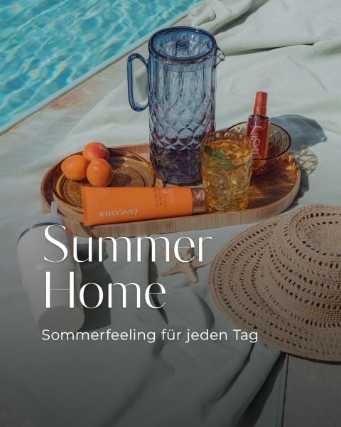 Eine Szene am Pool mit einem Strohhut, Sonnencreme, Aprikosen, einem Glaskrug, Getränken und einem Tablett. Der Text lautet: "Summer Home - Sommerfeeling für jeden Tag.