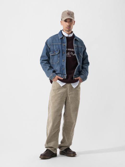 Ein Mann in Jeansjacke, Cordhose, karierter Mütze, Pullover und braunen Schuhen steht vor einem schlichten weißen Hintergrund.
