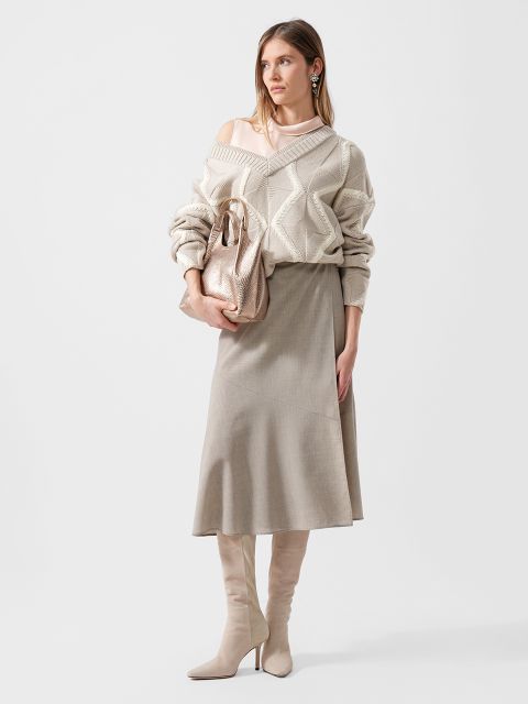 Frau in cremefarbenem Strickpullover, beigem Midirock, kniehohen Stiefeln und auffälligen Ohrringen, die eine Metallhandtasche vor einem schlichten Hintergrund halten.