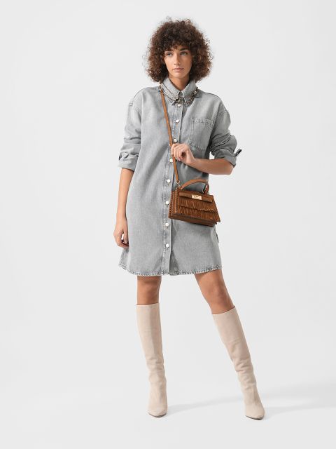 Eine Frau in einem hochgeknöpften Jeanshemd, beigefarbenen kniehohen Stiefeln und mit einer braunen Fransenhandtasche steht vor einem schlichten weißen Hintergrund.