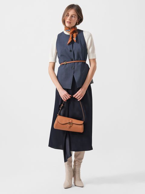 Frau mit blauer Weste, weißem, kurzärmeligem Oberteil, marineblauem Rock, beigen, kniehohen Stiefeln, brauner Handtasche und orangefarbenem Schal um den Hals.
