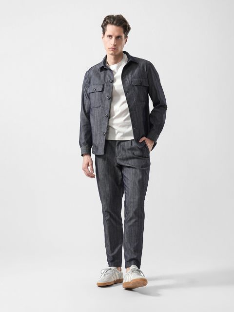 Mann in Freizeitkleidung: graue Button-up-Jacke und passende Hose, weißes T-Shirt und weiße Turnschuhe vor einem schlichten hellen Hintergrund.