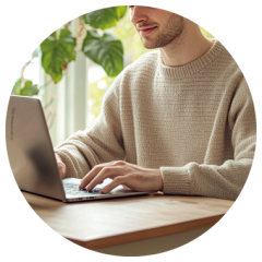 Mann im beigen Pullover, der an einem Holztisch auf einem Laptop tippt, mit Grünpflanzen und natürlichem Licht im Hintergrund.