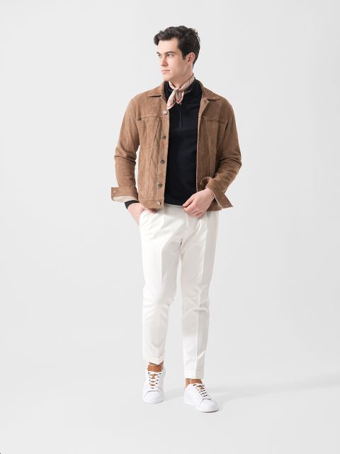 Ein Mann, der eine braune Wildlederjacke, ein schwarzes Hemd, eine weiße Hose, weiße Turnschuhe und einen Schal um den Hals trägt, steht vor einem schlichten weißen Hintergrund.