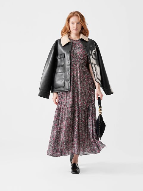 Frau in einem geblümten Maxikleid mit einer schwarzen Lederjacke über den Schultern, mit einer schwarzen Handtasche in der Hand und mit schwarzen Halbschuhen.