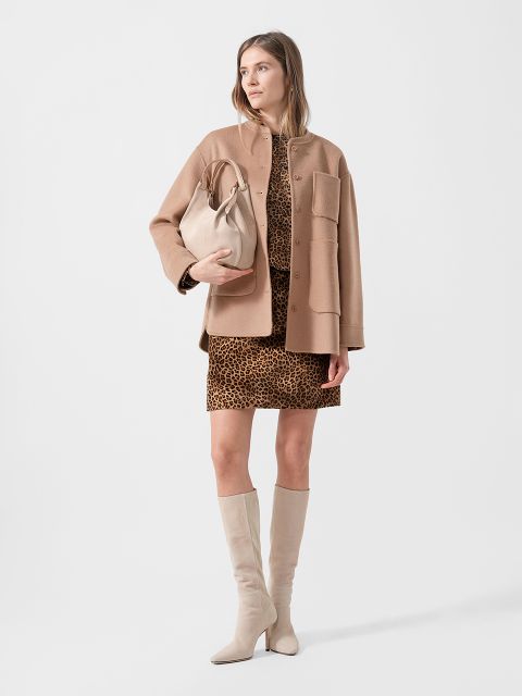 Eine Frau, die einen beigen Mantel über einem Kleid mit Leopardenmuster trägt, eine beigefarbene Tasche in der Hand hält und kniehohe beigefarbene Stiefel trägt, steht vor einem einfarbigen Hintergrund.