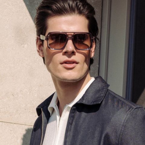 Ein Mann mit einer großen braunen Sonnenbrille und einer dunklen Jacke steht im Freien im Sonnenlicht und schaut leicht zur Seite.