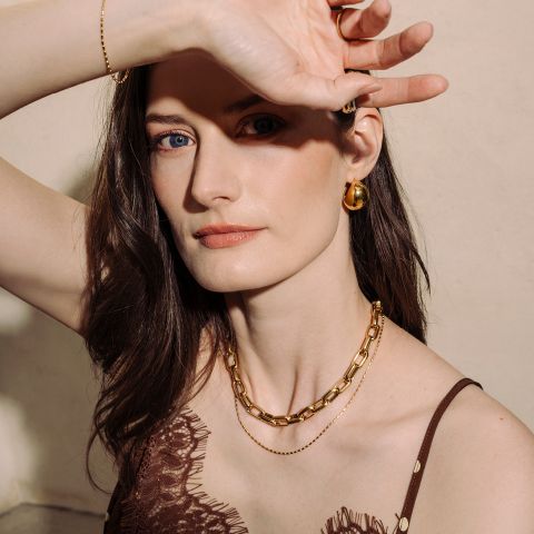 Eine Frau mit heller Haut und braunem Haar, die Goldschmuck trägt, posiert mit einem erhobenen Arm und blickt in die Kamera in weichem natürlichem Licht.