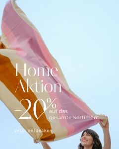 Lächelnde Frau, die eine große, bunte Decke gegen den Himmel hält; Text bietet 20 % Rabatt auf die gesamte Hauskollektion in deutscher Sprache.