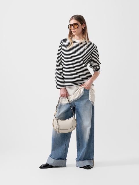 Frau in einem schwarz-weiß gestreiften Pullover, Jeans mit weitem Bein, Sonnenbrille und weißer Handtasche vor einem schlichten weißen Hintergrund.