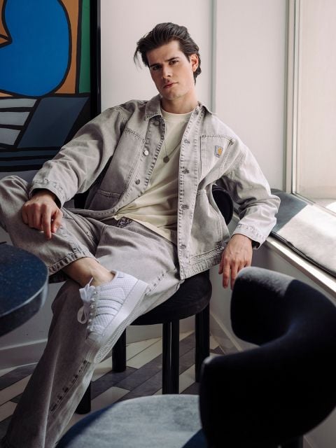 Ein junger Mann in legerer heller Jeans sitzt auf einem Stuhl am Fenster, ein Bein hochgelegt, in weißen Turnschuhen, in einem modernen, stilvollen Raum.