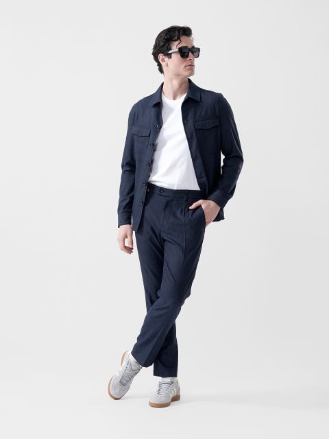 Ein Mann, der eine schwarze Sonnenbrille, eine marineblaue Jacke, ein weißes T-Shirt, eine marineblaue Hose und graue Turnschuhe trägt, steht mit einem Fuß vor dem anderen gekreuzt und schaut zur Seite.