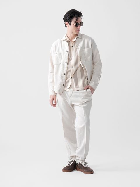 Ein Mann mit Sonnenbrille, hellbeiger Jacke, gestreiftem Hemd und passender Hose steht mit den Händen in den Taschen vor einem schlichten weißen Hintergrund.