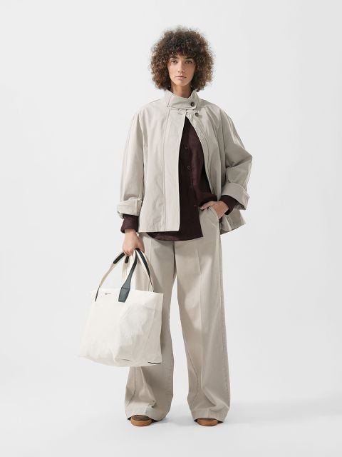 Eine Frau mit lockigem Haar trägt eine beigefarbene Jacke und eine Hose mit weitem Bein, ein dunkles Hemd, braune Schuhe und hat eine große weiße Tragetasche dabei.