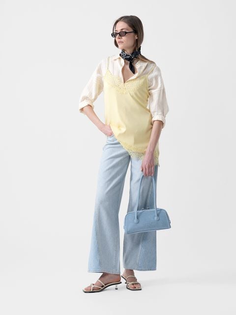 Frau mit Sonnenbrille, die ein gelbes Unterhemd über einem gestreiften Hemd, hellblaue Jeans mit weitem Bein, Sandalen und ein Halstuch trägt und eine blaue Handtasche hält.