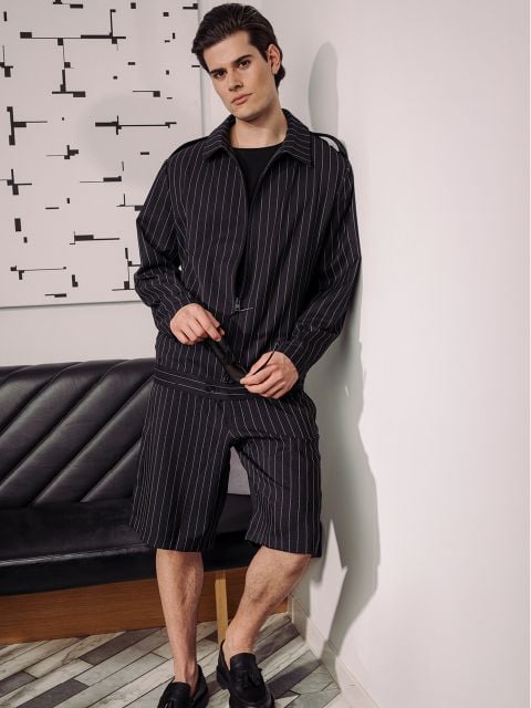 Ein Mann in schwarzer Nadelstreifenjacke und Shorts steht an einer Wand, neben einem schwarzen Sofa, hinter dem abstrakte Kunst hängt.