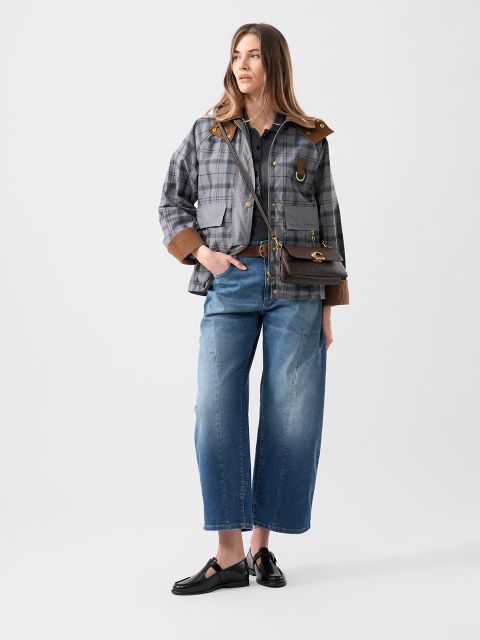 Eine Frau in einer karierten Jacke, blauen Jeans mit weitem Bein, schwarzen Schuhen und einer braunen Umhängetasche steht vor einem schlichten weißen Hintergrund.