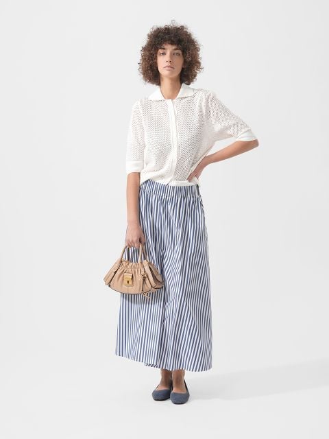 Frau in einem weißen kurzärmeligen Strickoberteil, einem blau-weiß gestreiften Maxirock, marineblauen Flats und einer beigen Handtasche vor einem schlichten Hintergrund.