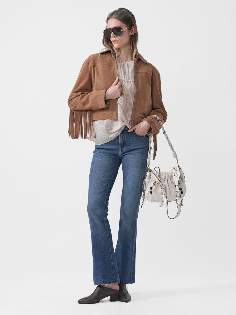 Eine Frau mit Sonnenbrille trägt eine braune Fransenjacke, ein beiges Oberteil, blaue Jeans, schwarze Schuhe und eine weiße Handtasche und posiert vor einem weißen Hintergrund.