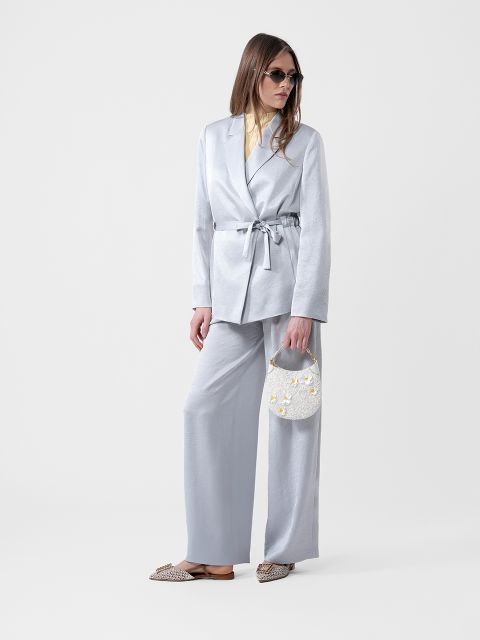 Frau in einem hellgrauen Anzug mit Gürtelblazer, gelbem Hemd, Sonnenbrille, einer kleinen weißen Handtasche und gemusterten Flats.