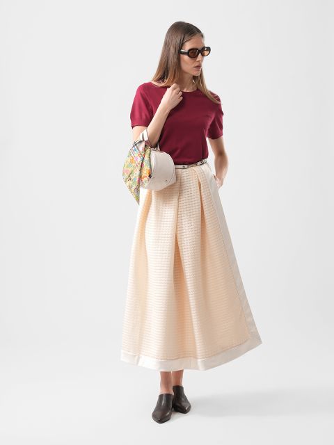 Frau mit rotem Oberteil, cremefarbenem langem Rock, schwarzen Schuhen, Sonnenbrille und einer gemusterten Handtasche in der Hand, vor einem schlichten Hintergrund stehend.