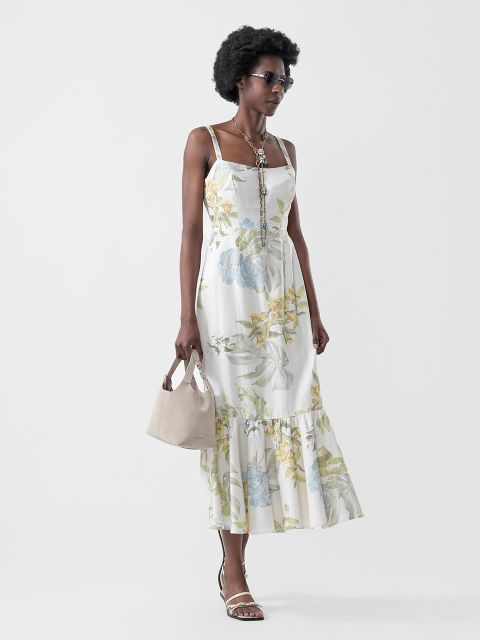 Eine Frau in einem geblümten weißen Sonnenkleid, Sandalen, Sonnenbrille und Halskette, die eine beigefarbene Handtasche trägt, posiert vor einem schlichten hellen Hintergrund.