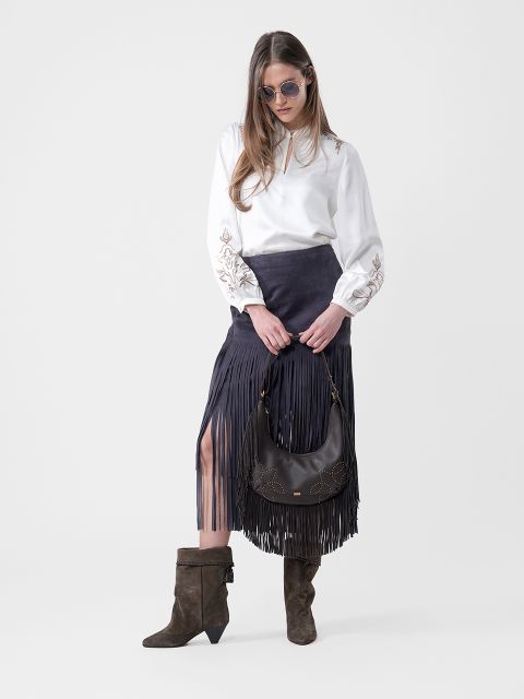 Frau mit Sonnenbrille, weißer Bluse, schwarzem Fransenrock, braunen Stiefeln und einer schwarzen Handtasche in der Hand, vor einem schlichten Hintergrund.