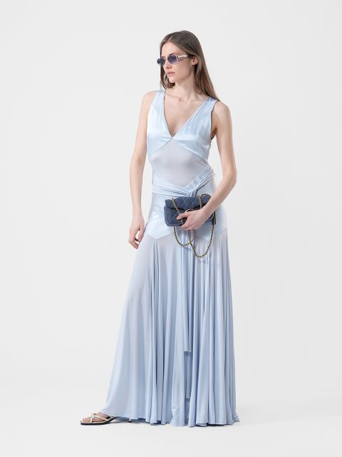 Frau in einem hellblauen, fließenden, ärmellosen Kleid mit Sonnenbrille, die eine dunkle Handtasche mit Kette in der Hand hält, stehend vor einem schlichten weißen Hintergrund.