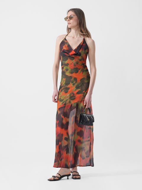 Frau, die ein ärmelloses, orange und grün gemustertes Maxikleid, Sandalen und eine Sonnenbrille trägt und eine kleine schwarze Handtasche in der Hand hält, steht vor einem weißen Hintergrund.