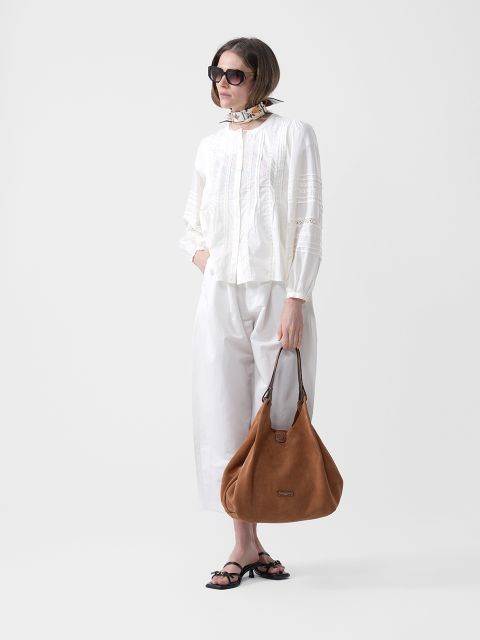 Frau mit Sonnenbrille, weißer Bluse und Hose, einem Schal um den Hals, schwarzen Sandalen und einer großen braunen Wildlederhandtasche in der Hand.
