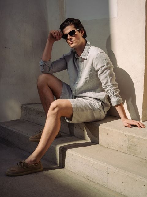 Ein Mann mit Sonnenbrille, hellem Leinenhemd und kurzen Hosen sitzt auf einer Betontreppe im Sonnenlicht und stützt einen Arm auf sein Knie.