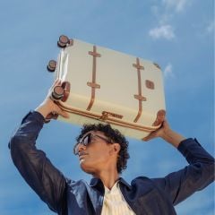 Eine Person mit Sonnenbrille und dunkler Jacke hält einen cremefarbenen Koffer mit braunen Riemen über ihren Kopf vor einem blauen Himmel.