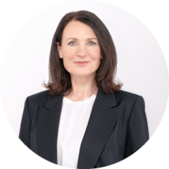 Frau mit schulterlangen braunen Haaren, die einen schwarzen Blazer und eine weiße Bluse trägt, lächelt sanft vor einem schlichten hellen Hintergrund.