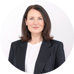 Frau mit schulterlangen braunen Haaren, die einen schwarzen Blazer und eine weiße Bluse trägt, lächelt sanft vor einem schlichten hellen Hintergrund.