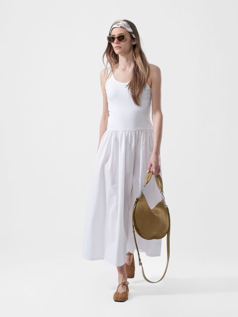 Frau in einem weißen ärmellosen Maxikleid, Sonnenbrille, Kopftuch, hellbraunen Flats und mit einer hellbraunen runden Handtasche vor einem schlichten weißen Hintergrund.
