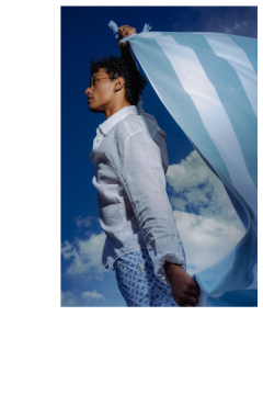 Eine Person in einem weißen Hemd und gemusterten Shorts hält ein blau-weiß gestreiftes Tuch vor einen blauen Himmel mit Wolken.
