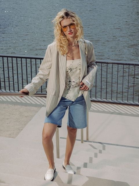 Eine Frau, die eine Sonnenbrille, einen gestreiften Blazer, ein geblümtes Oberteil, Jeansshorts und weiße Schuhe trägt, steht an einer Uferpromenade mit einem Geländer hinter ihr.
