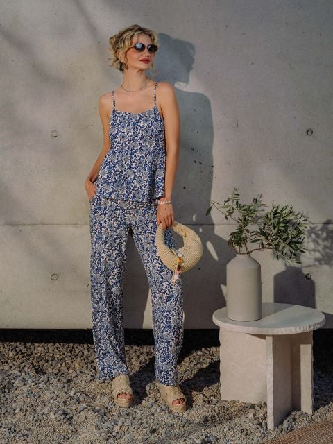Frau in blauem, geblümtem Oberteil und passender Hose, die einen Strohhut trägt, steht an einem Tisch mit einer Vase und einem Olivenzweig vor einer Betonwand.