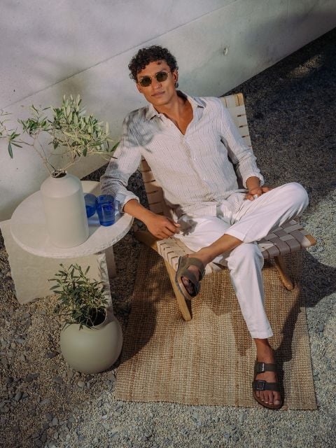 Ein Mann mit Sonnenbrille und gestreiftem Hemd macht es sich auf einem geflochtenen Stuhl neben einem runden Tisch mit Pflanzen und blauen Gläsern auf einer strukturierten Matte im Freien bequem.