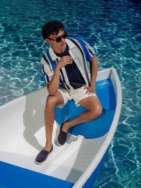 Ein Mann mit Sonnenbrille und gestreiftem Hemd sitzt auf einem kleinen weiß-blauen Boot in einem Pool mit klarem, glitzerndem Wasser.