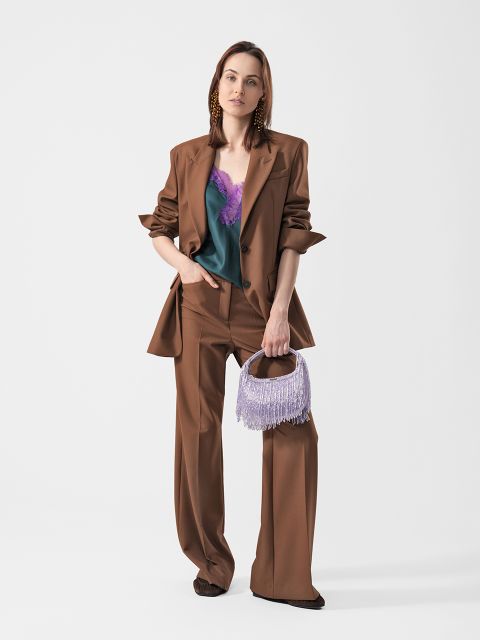 Eine Frau in einem braunen Anzug mit weiter Hose und einem blaugrünen Oberteil mit violetter Spitze, die eine lavendelfarbene Tasche mit Fransen in der Hand hält und vor einem schlichten weißen Hintergrund steht.
