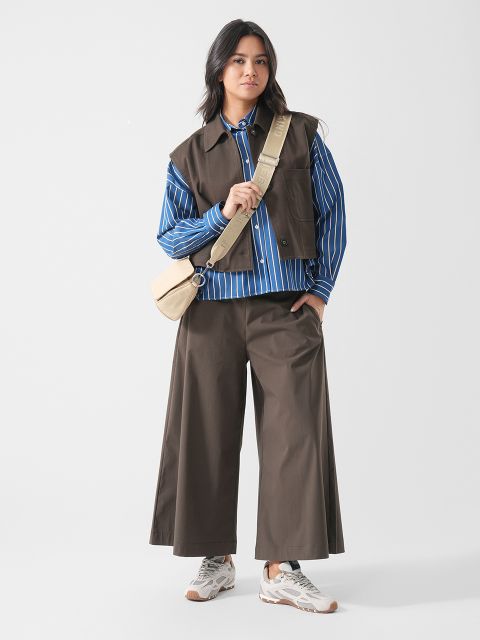 Eine Frau in einer weiten braunen Hose, einer braunen Weste, einem blau gestreiften Hemd, weißen Sneakers und mit einer beigen Umhängetasche steht vor einem schlichten weißen Hintergrund.