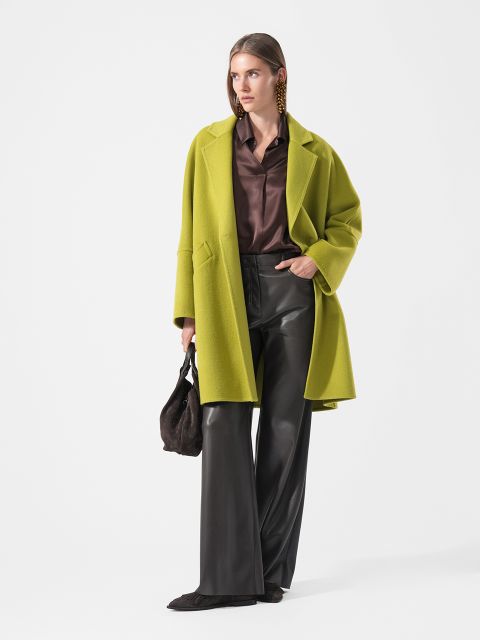 Eine Frau in einem limettengrünen Mantel, einem braunen Hemd, einer schwarzen Lederhose und schwarzen Schuhen, die eine braune Tasche in der Hand hält und vor einem weißen Hintergrund steht.