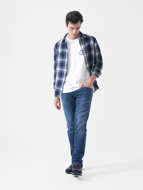 Ein Mann in einem blauen Karohemd, einem weißen T-Shirt, blauen Jeans und Turnschuhen, der vor einem schlichten weißen Hintergrund geht und nach unten schaut.