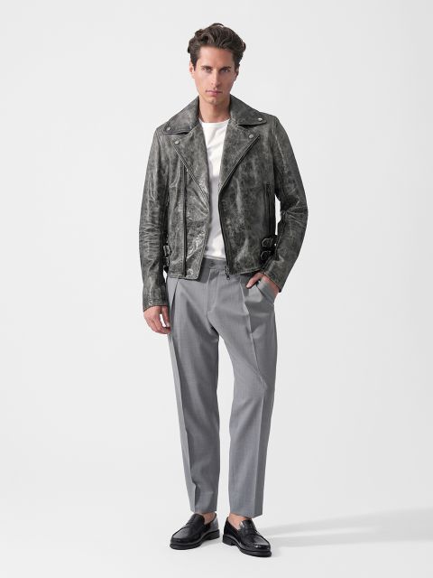 Eine Person, die eine graue Lederjacke, ein weißes Hemd, eine graue Hose und schwarze Slipper trägt, steht vor einem schlichten weißen Hintergrund.