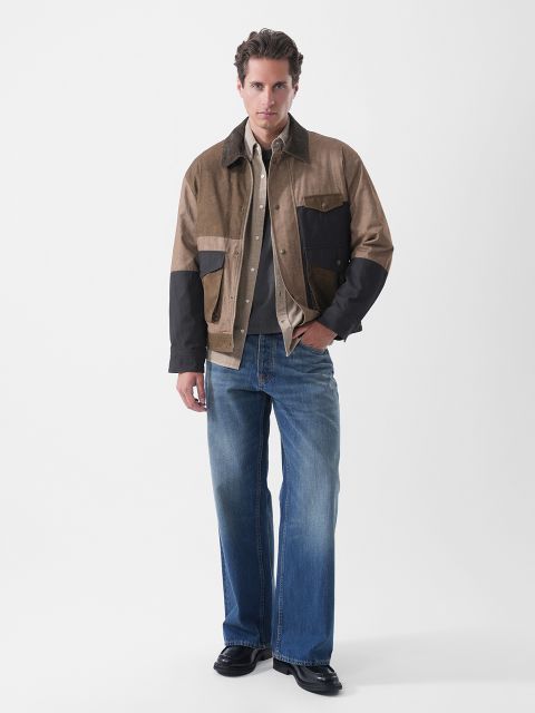 Ein Mann steht in Freizeitkleidung da: eine Jacke in Braun und Schwarz im Color-Blocking-Stil, ein helles Hemd, blaue Jeans und schwarze Schuhe; sein Gesichtsausdruck ist neutral, vor einem schlichten Hintergrund.