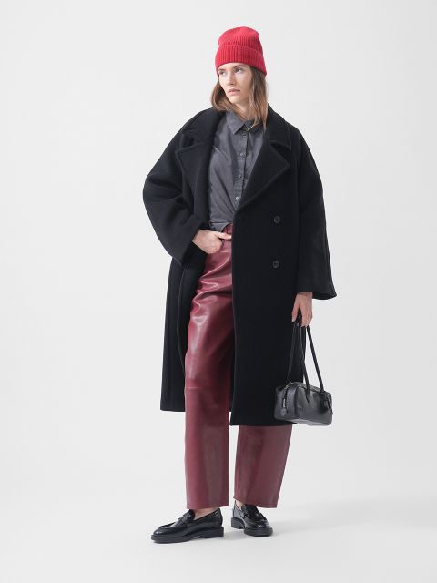 Eine Frau mit roter Mütze, schwarzem Mantel, grauem Shirt, roter Lederhose und schwarzen Loafern hält eine schwarze Handtasche in der Hand und posiert vor einem weißen Hintergrund.