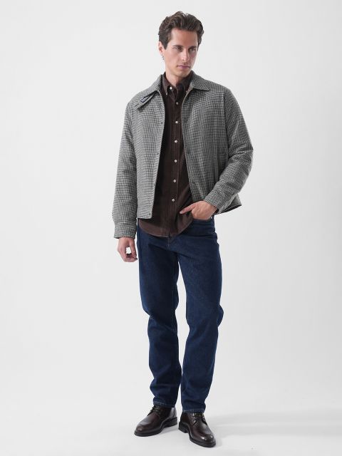 Ein Mann in einer grau karierten Jacke, einem braunen Hemd, blauen Jeans und braunen Schuhen steht vor einem schlichten weißen Hintergrund.